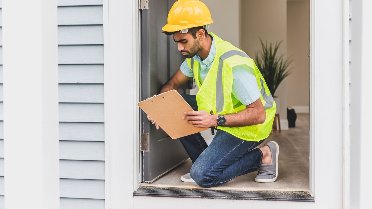An inspector in hi-vis checking the base of an exterior wall, clipboard in hand